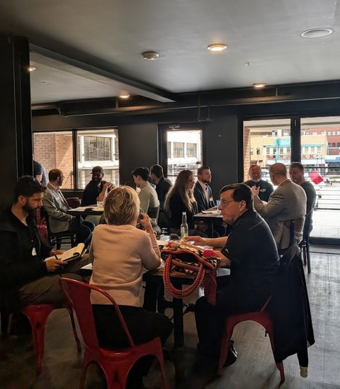 Small groups of members at tables in a bright room with tall windows