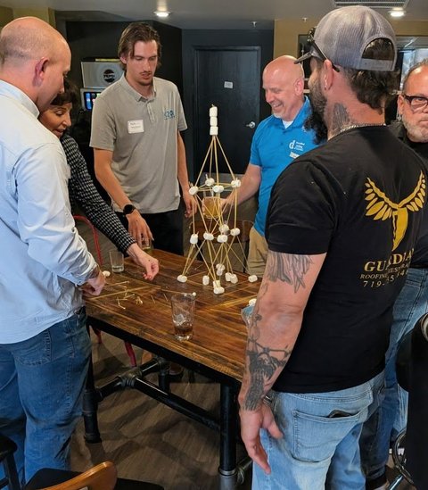 Members standing around a spaghetti and marshmallow tower during a team challenge